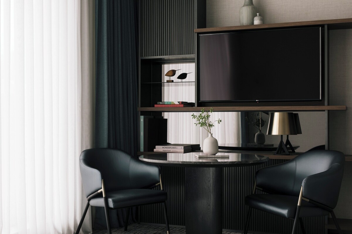Une pièce moderne avec une table ronde, deux chaises noires, un téléviseur à écran plat posé sur une étagère en bois, un vase avec une plante et des livres sur une étagère inférieure.