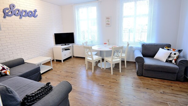 Una sala de estar moderna con pared de ladrillo blanco, un televisor de pantalla plana, sofás grises, una mesa de comedor con sillas y ventanas grandes.