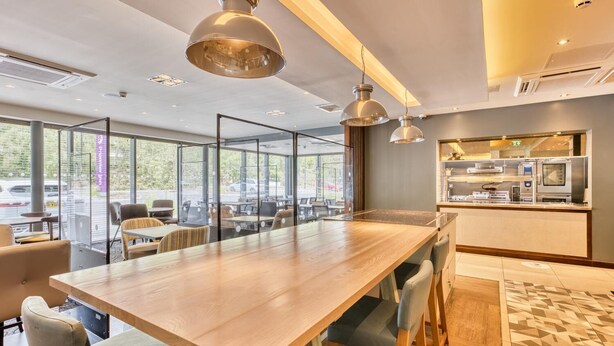 A modern conference room with a long table, chairs, pendant lighting, and a kitchen area visible through glass partitions.