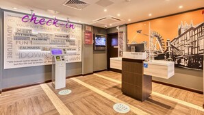 A hotel reception area with a check-in kiosk, a digital screen, and a wall with various travel-related words.