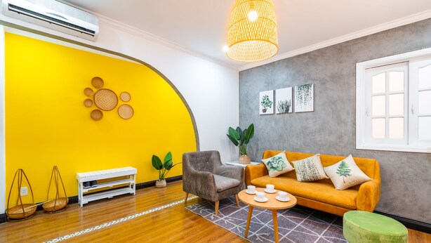 A modern living room with a yellow accent wall, a grey armchair, a yellow sofa, and a green ottoman. There is a white bench, a small table with cups, and a hanging light fixture. - Chin Stays - Hotel and Apartment (Da Nang)