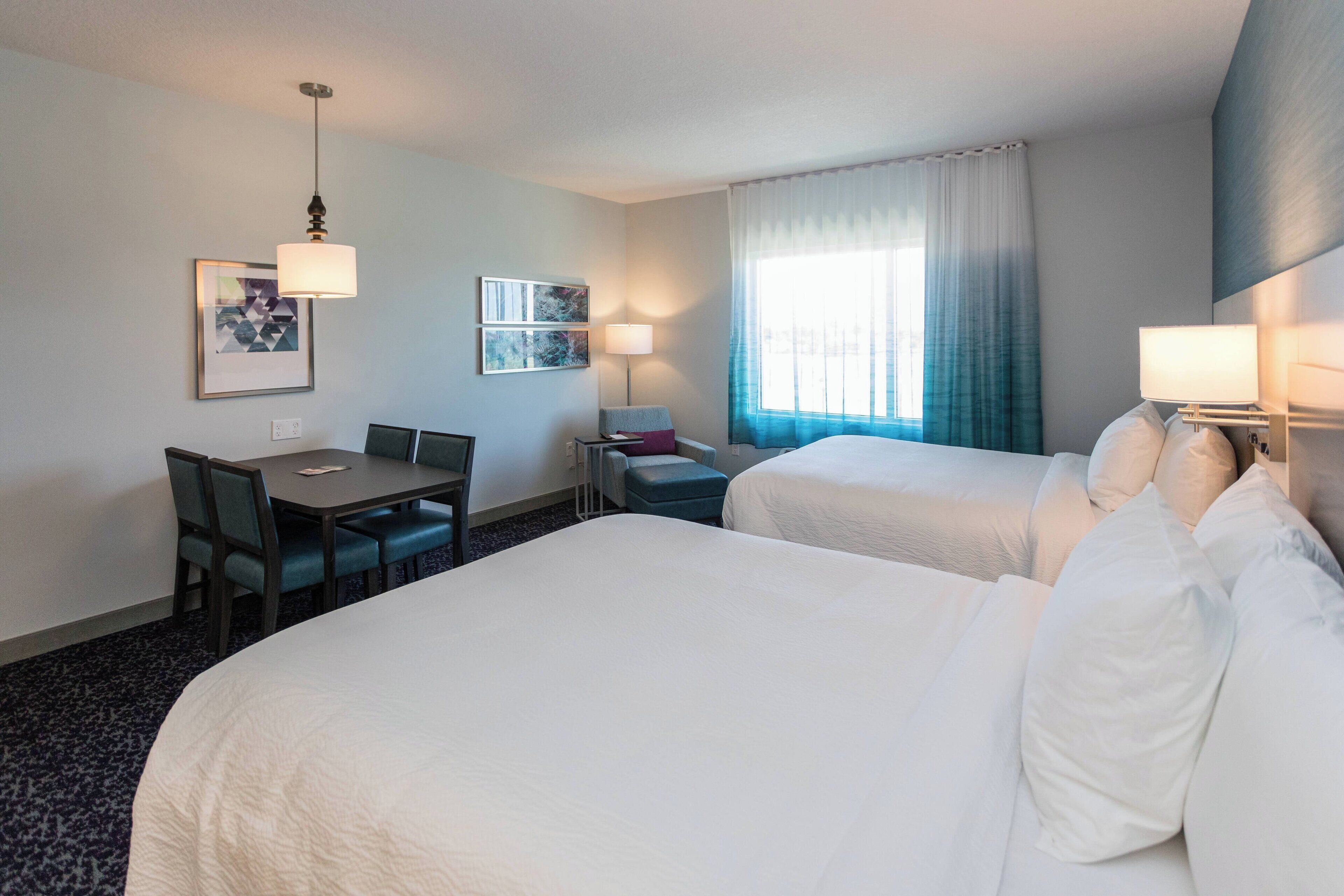 Quarto de hotel com duas camas, uma escrivaninha, cadeiras, luminária e janela com cortinas.