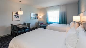 Quarto de hotel com duas camas, uma escrivaninha, cadeiras, luminária e janela com cortinas.