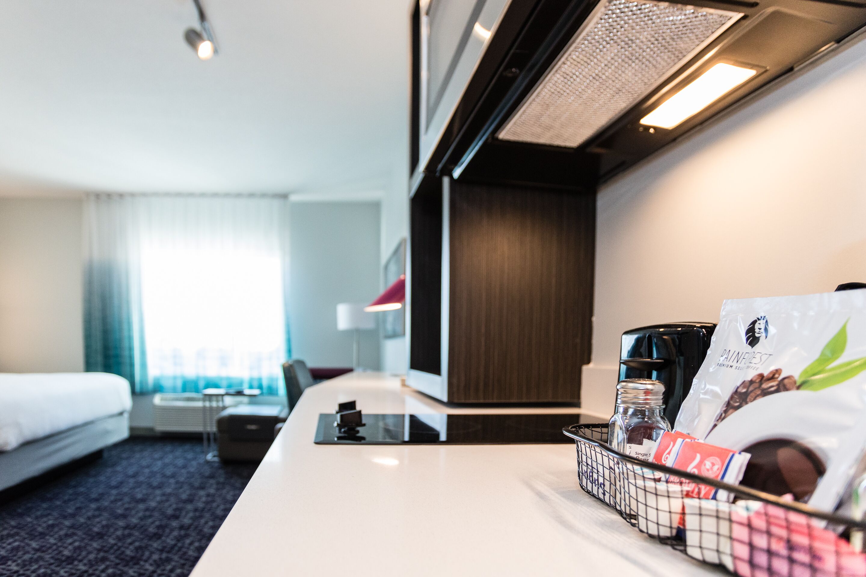 A hotel room with a bed, a desk with a coffee maker, a basket of toiletries, and a window with curtains.