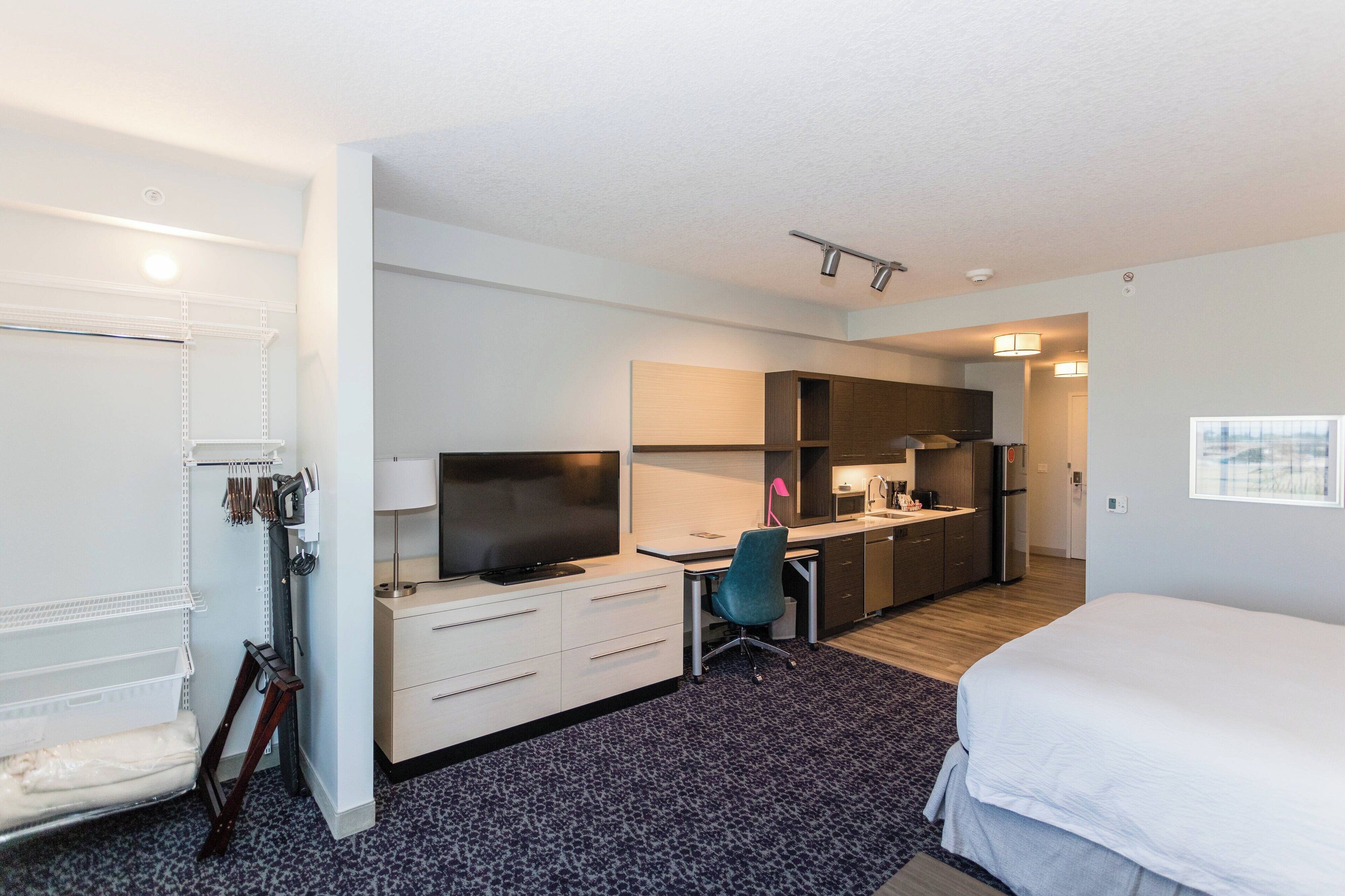 A hotel room with a bed, a desk with a computer, a chair, a TV, and a kitchenette.