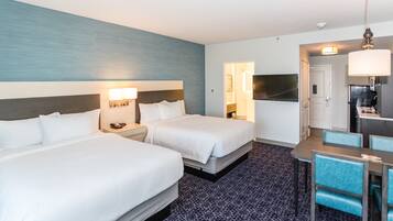 A hotel room with two beds, a desk, a chair, a TV, and a kitchenette.