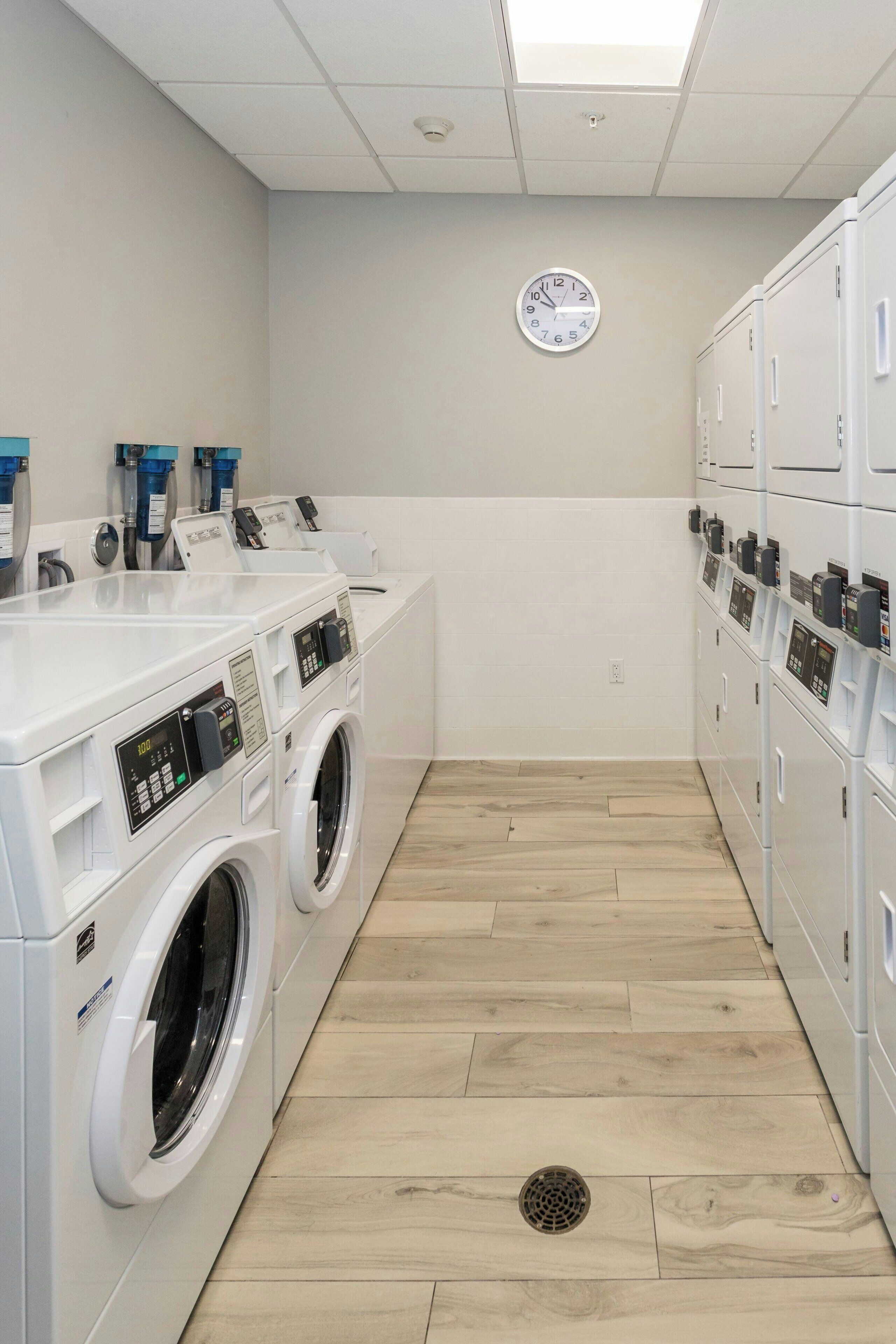 A laundry facility with multiple washing machines and dryers, a clock on the wall, and a wooden floor.