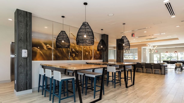 A modern bar area with wooden tables and blue chairs, featuring black pendant lights and a large abstract mural on the wall. - TownePlace Suites by Marriott Port St. Lucie I-95 (Port Saint Lucie)