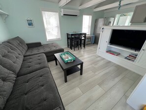A living room with a grey sectional sofa, a small black table with a green tray, a TV on a white stand, and a dining area with a table and chairs.