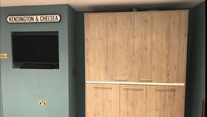 Une chambre dotée d’une grande armoire, d’un téléviseur à écran plat fixé au mur et d’une enseigne au-dessus indiquant « Kensington & Chelsea ».