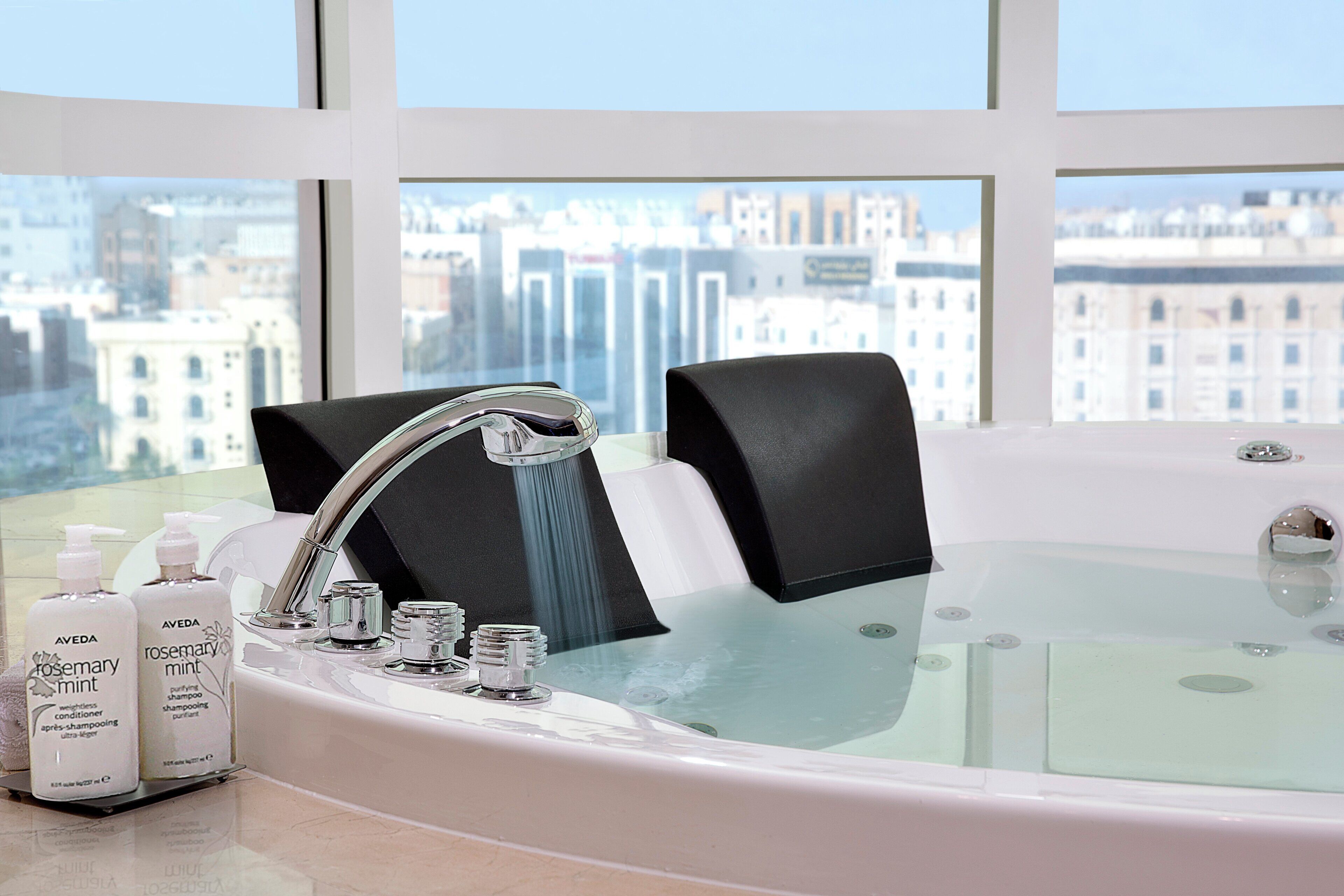 A bathroom with a bathtub, two bottles of shampoo, and a city view through large windows.