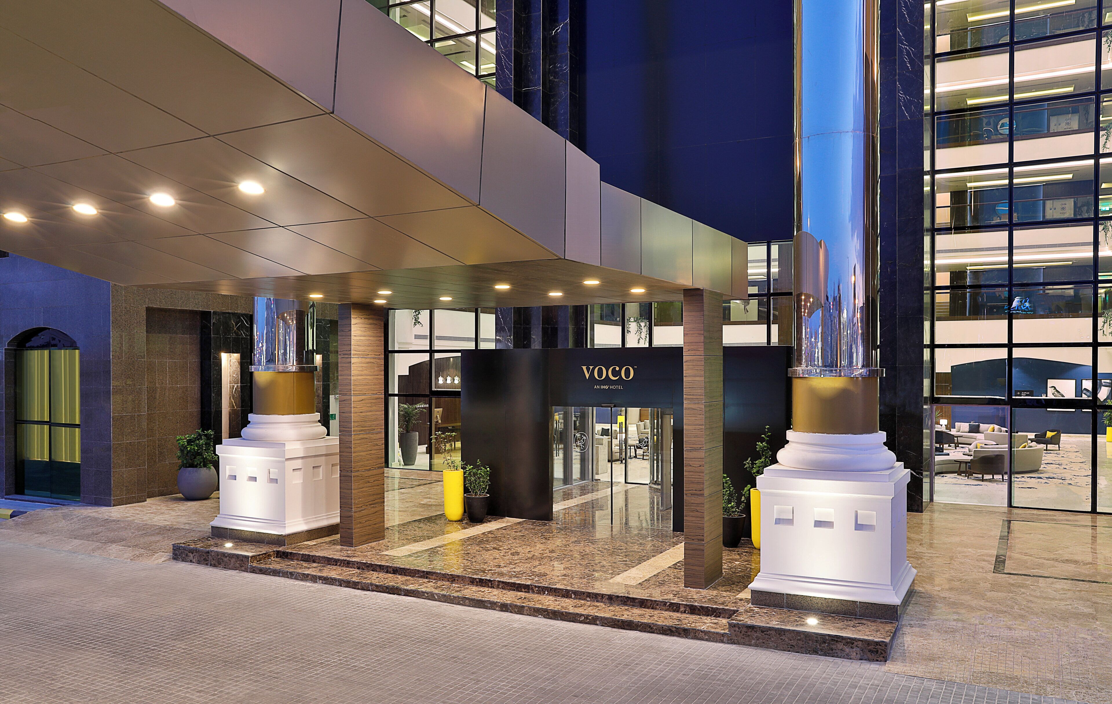 Entrada de hotel com design moderno, destacando fachada de vidro, balcão de recepção e área de estar com sofás.