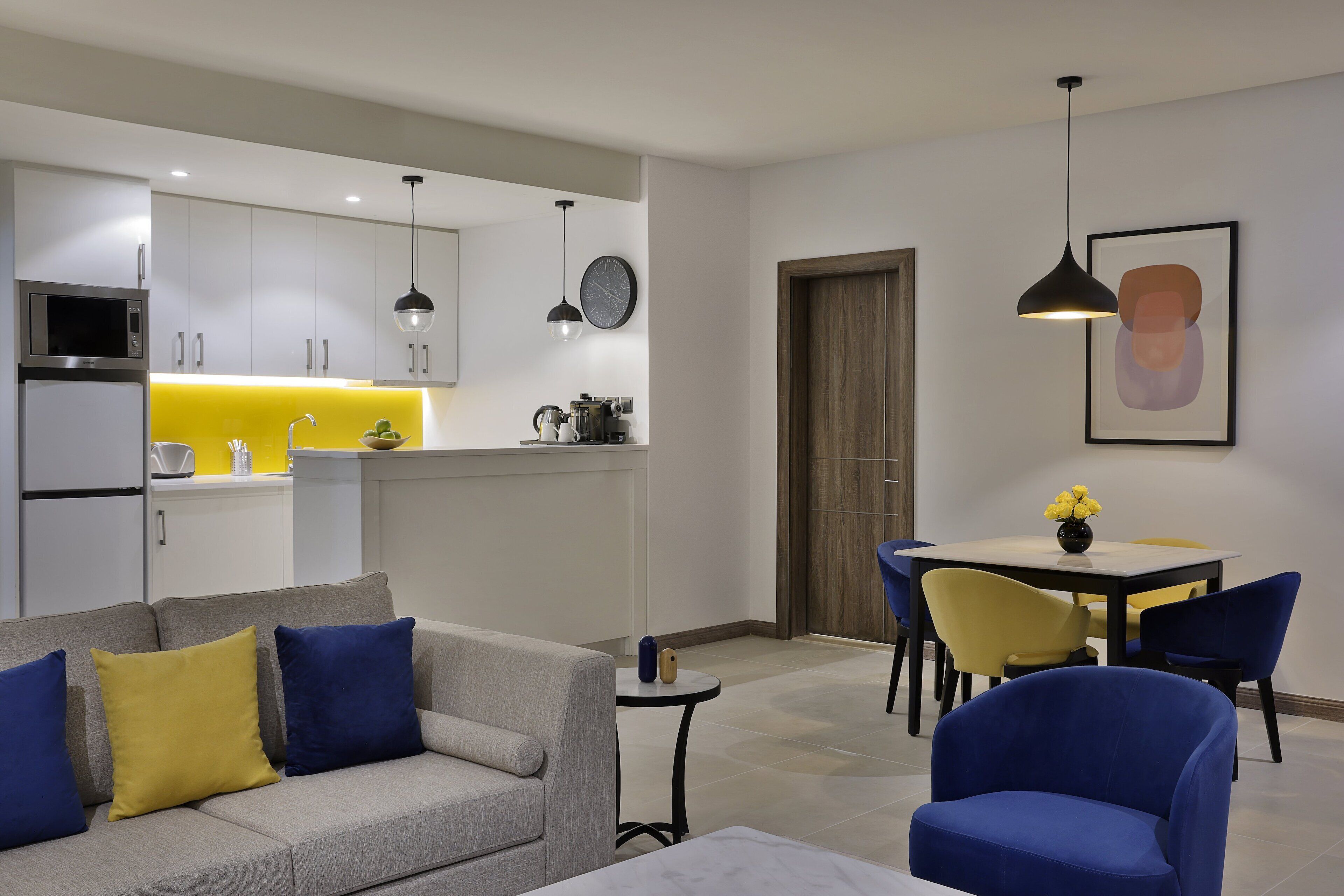 Una sala moderna con un sofá gris, sillas azules y amarillas, una mesita y una zona de comedor con una mesa y sillas amarillas.
