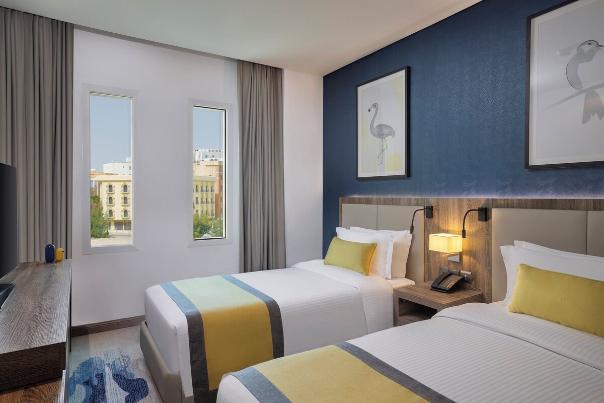 A hotel room with two beds, a desk, a nightstand with a lamp, and a view of buildings through the windows.