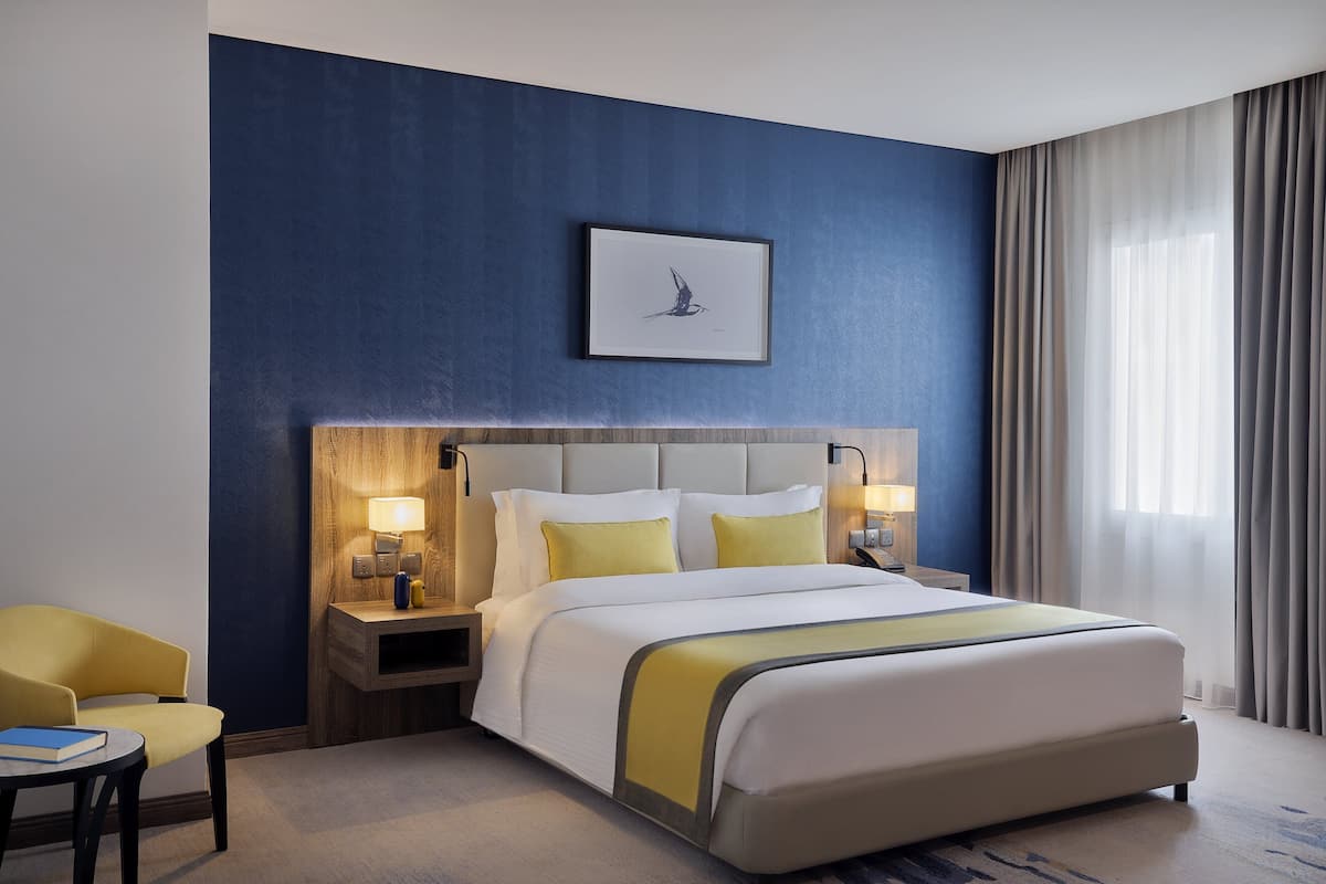 A hotel room with a large bed, a bedside table with a lamp, a chair, and a framed picture on the wall.