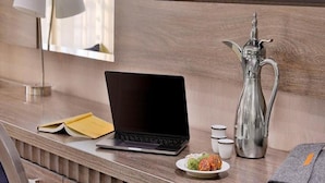 A hotel room with a wooden desk, a laptop, a silver pitcher, a plate with food, a book, and a TV mounted on the wall.