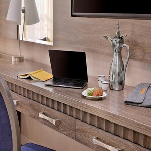 A hotel room with a wooden desk, a laptop, a silver pitcher, a plate with food, a book, and a TV mounted on the wall. - voco Al Khobar by IHG (Al Khobar)