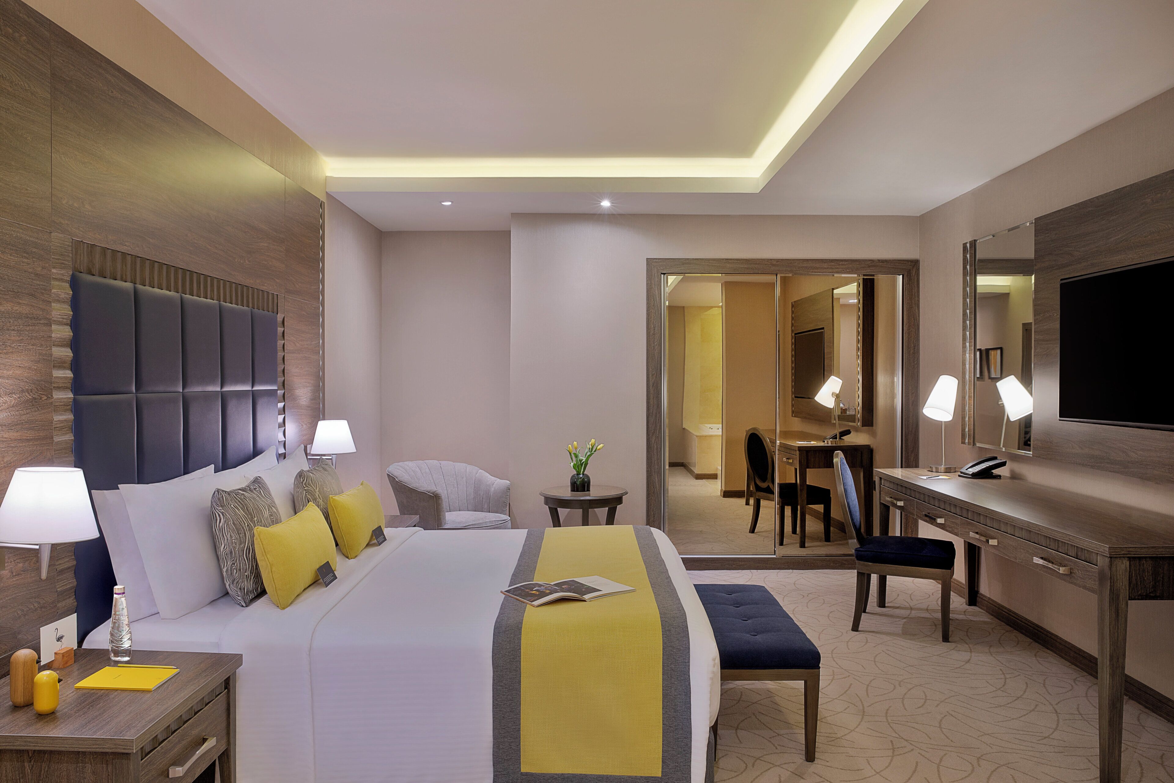 Une chambre d’hôtel avec un lit, des tables de chevet, un bureau, une chaise, une télévision et une salle de bain visible à travers une porte ouverte.