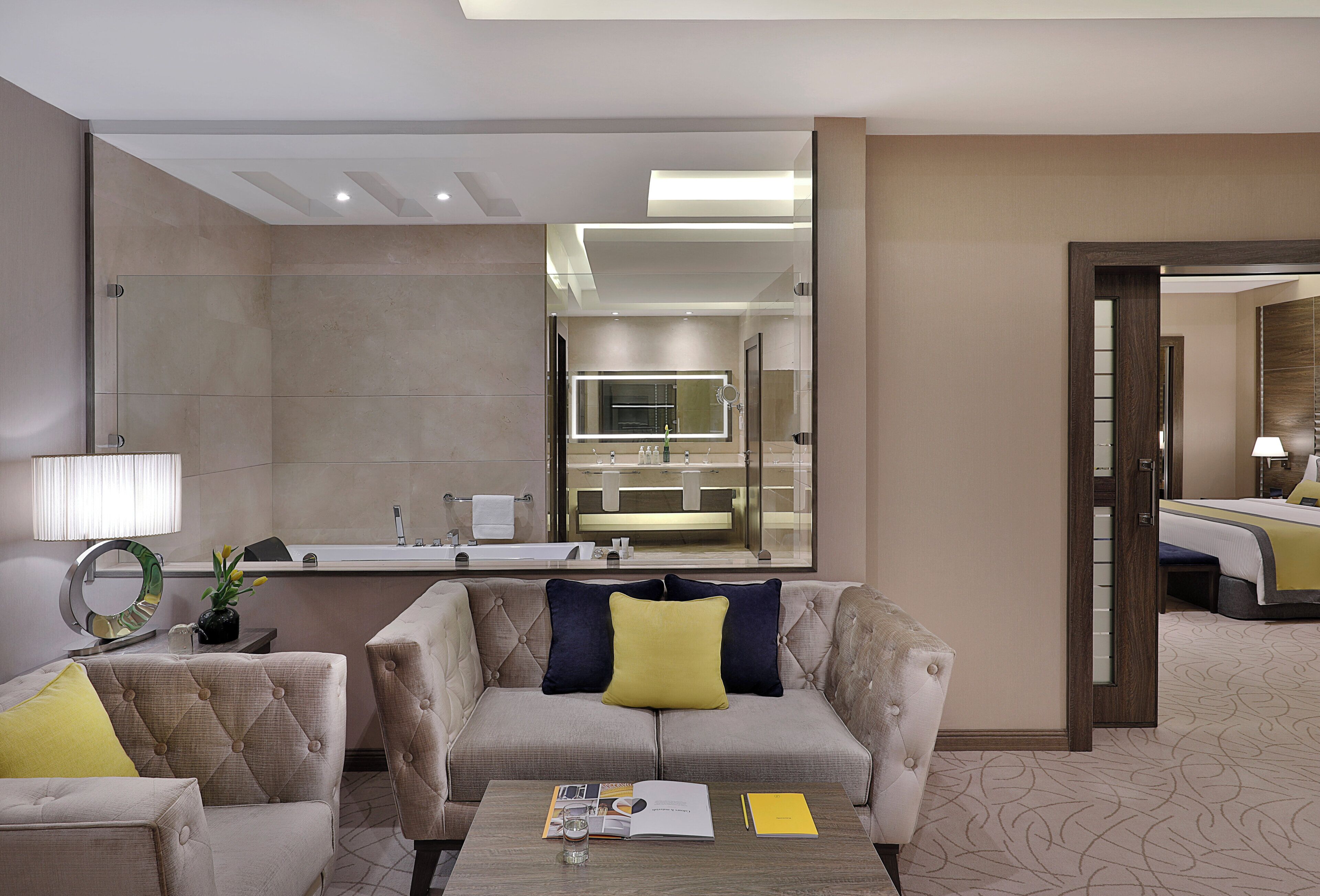 A hotel room with a sofa, a coffee table with a magazine and a glass, a bathroom with a mirror and sink, and a bed with a lamp.