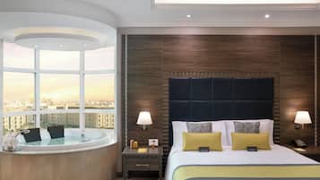 A hotel room with a large bed, two bedside lamps, a view of the city through a large window, and a bathtub.
