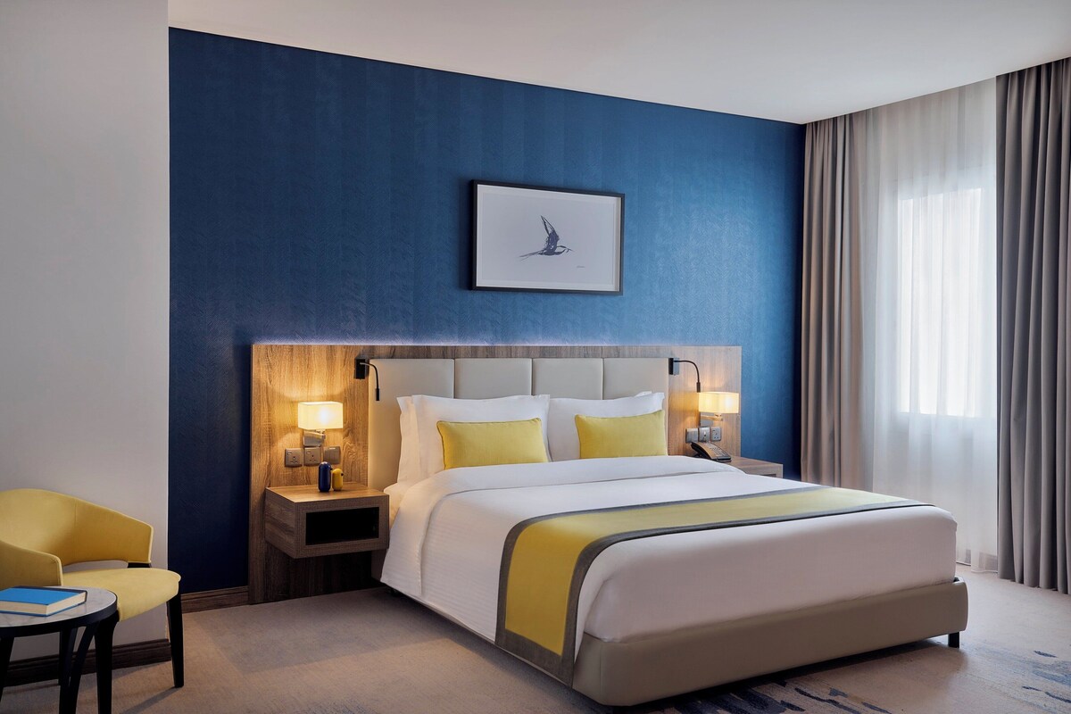 A hotel room with a large bed, a bedside table with a lamp, a chair, and a framed picture on the wall.