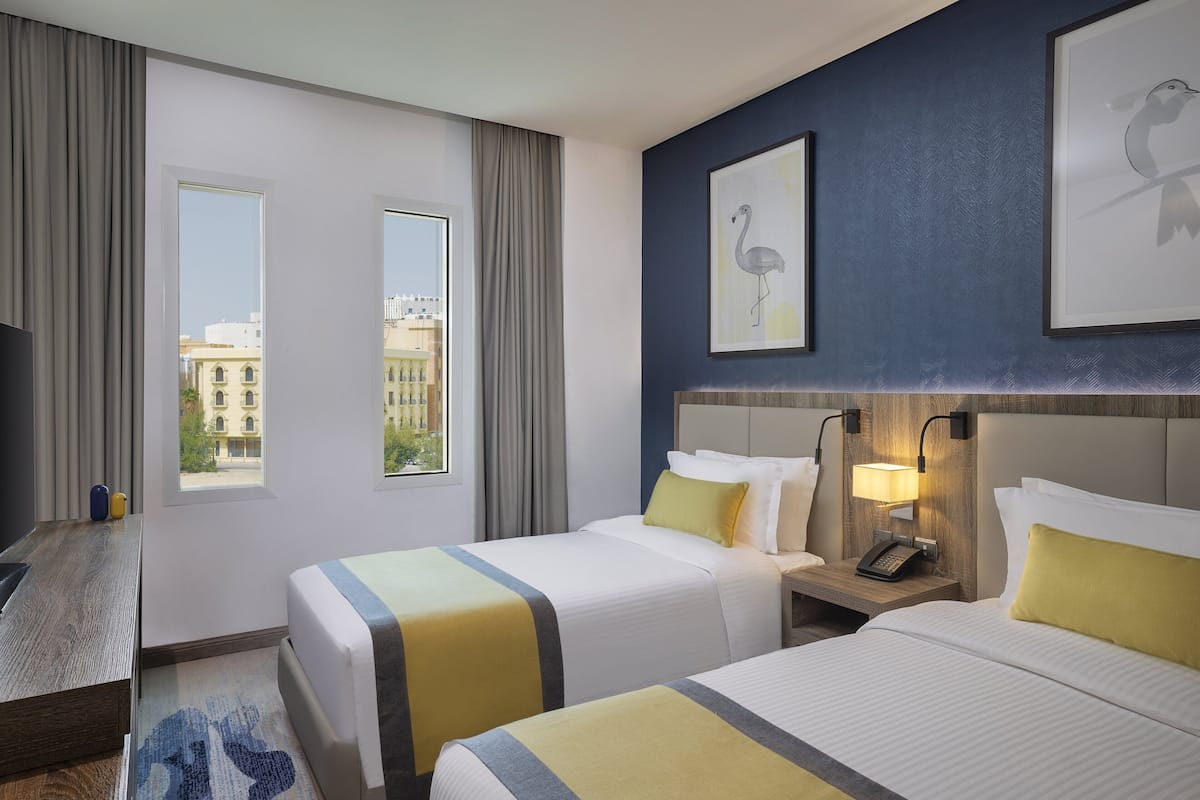 A hotel room with two beds, a desk, a nightstand with a lamp, and a view of buildings through the windows.