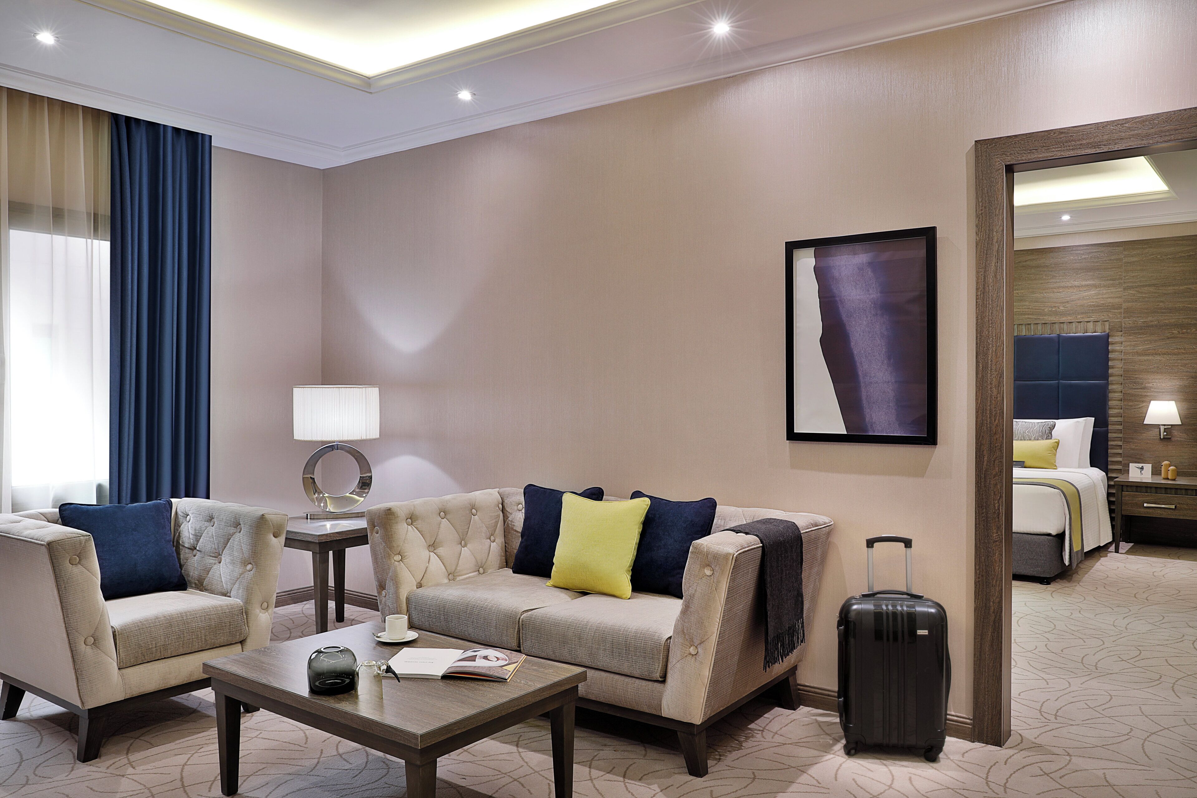 A hotel room with a sofa, armchairs, a coffee table, a lamp, a bed, a nightstand, a mirror, and a suitcase.