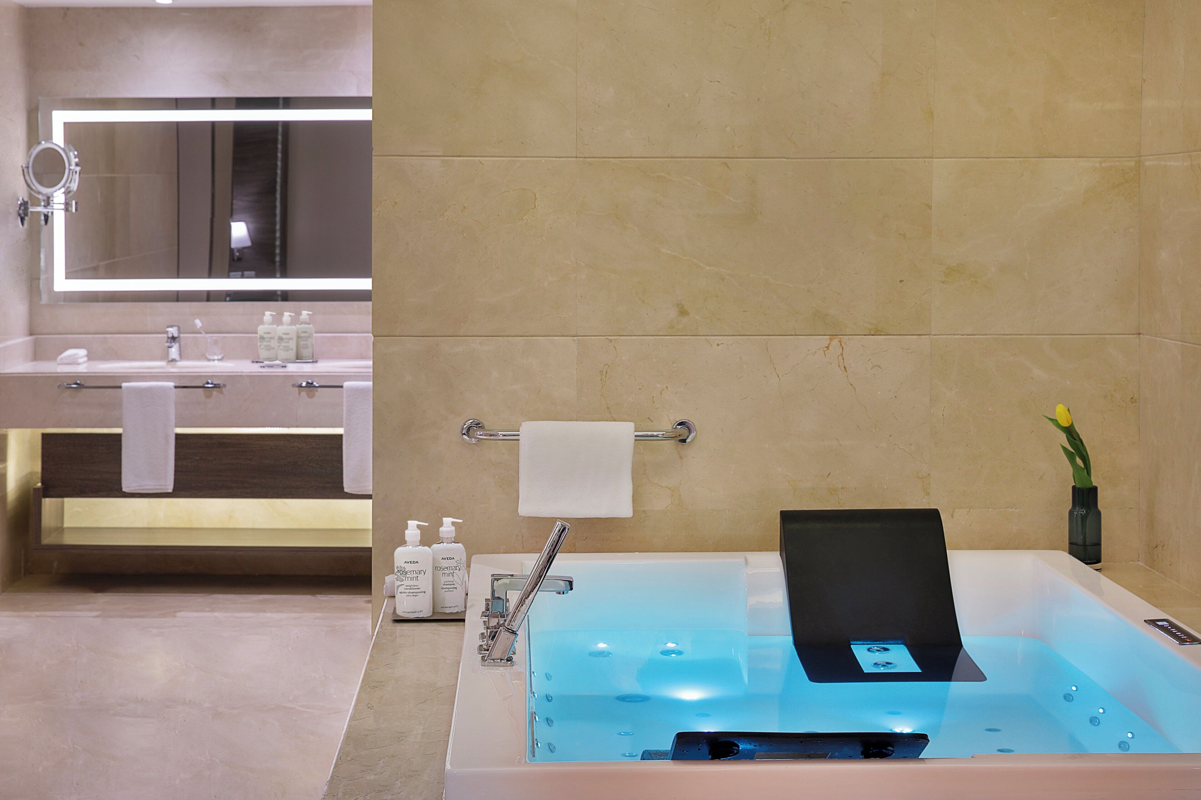 A modern bathroom with a large mirror, a sink with toiletries, and a bathtub with a control panel.