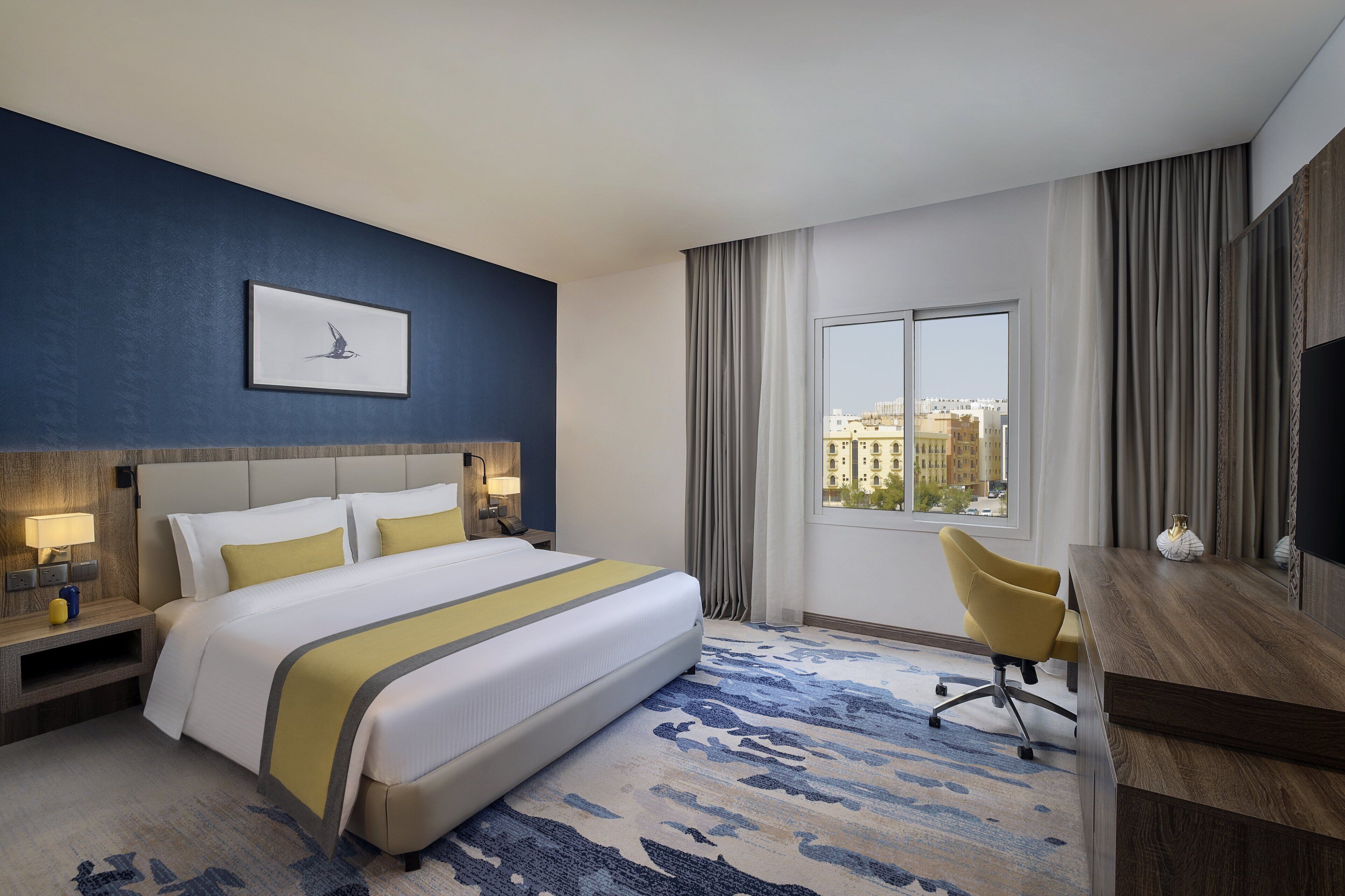 A hotel room with a large bed, a desk, a chair, a TV, and a window with a city view.