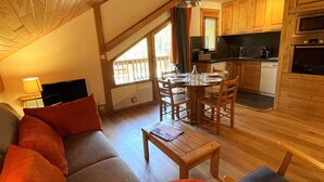 A cozy living room with a sofa, a coffee table, and a dining area with a table and chairs. A kitchen with wooden cabinets and appliances is visible in the background. - Renovated 2-room apartment, 5 people, Méribel Altiport, ski-in/ski-out (Méribel)