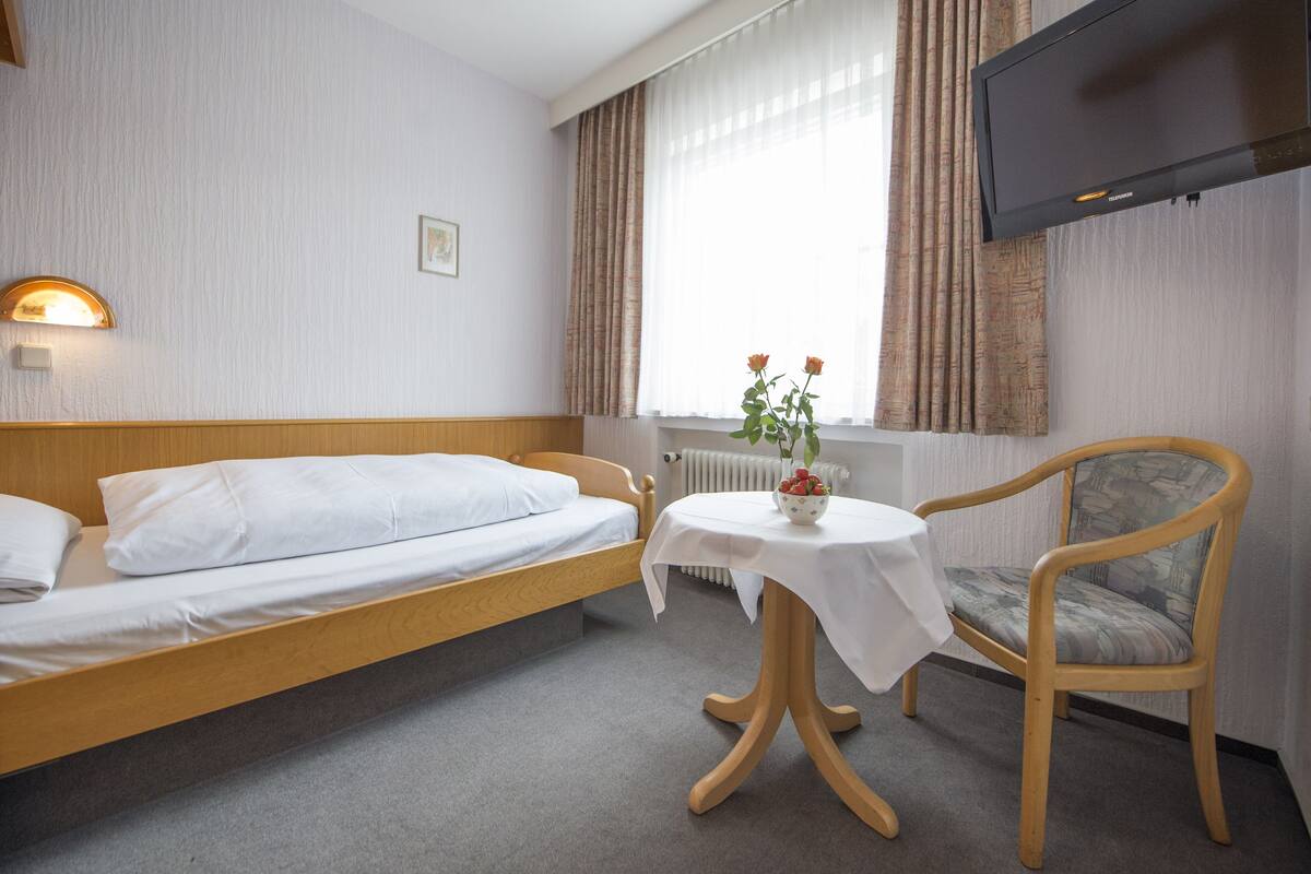 Habitación de hotel con una cama individual, una mesita con un jarrón de flores, una silla y un televisor montado en la pared.