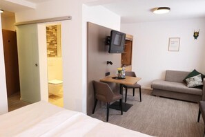 A hotel room with a bed, a small dining table with chairs, a television mounted on the wall, a sofa, and a framed picture on the wall.