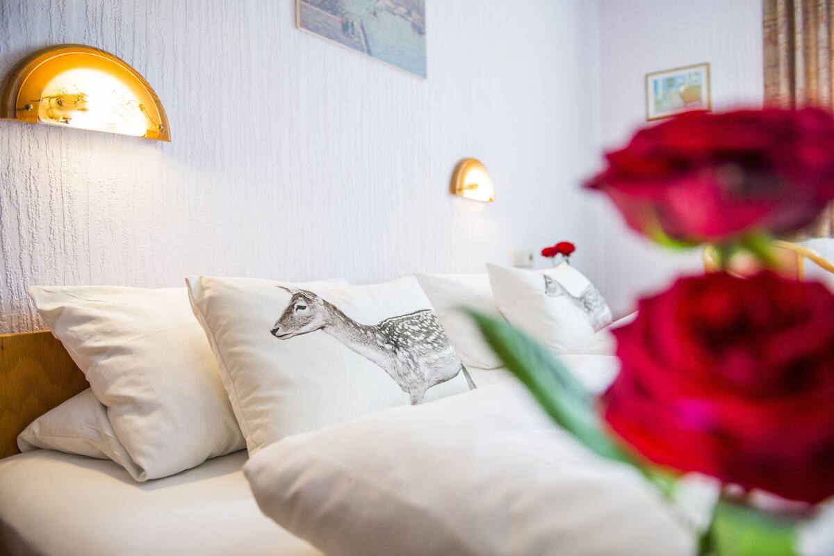 Una cama con ropa de cama blanca y un cojín con diseño de ciervo, una lámpara de pared y una rosa roja en primer plano.