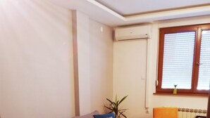 A living room with a grey sofa, a small white table, and an orange chair. There is a window with blinds and an air conditioning unit on the wall.