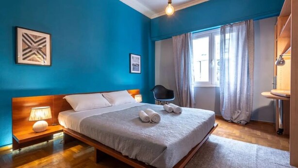 A bedroom with a large bed, a bedside table with a lamp, a chair, a window with curtains, and a wall-mounted picture. - S&K Polyxene Suite in Center of Athens (Athens)