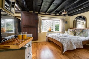 A bedroom with a wooden floor, a bed with a patterned bedspread, a window with a view of greenery, and a wooden cabinet with drawers. - Forest Drove Cottage (Ringwood)