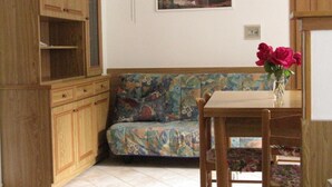 Ein Zimmer mit einem Holzkasten, einem Sofa mit farbenfrohem Blumenmuster, einem Landschaftsbild und einer Vase mit roten Blumen auf einem Tisch.