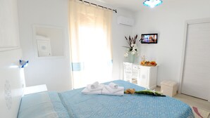 A neatly arranged bedroom with a bed, a small white cabinet, a television, and a window with curtains. - INAH Boutique Rooms (Dorgali)