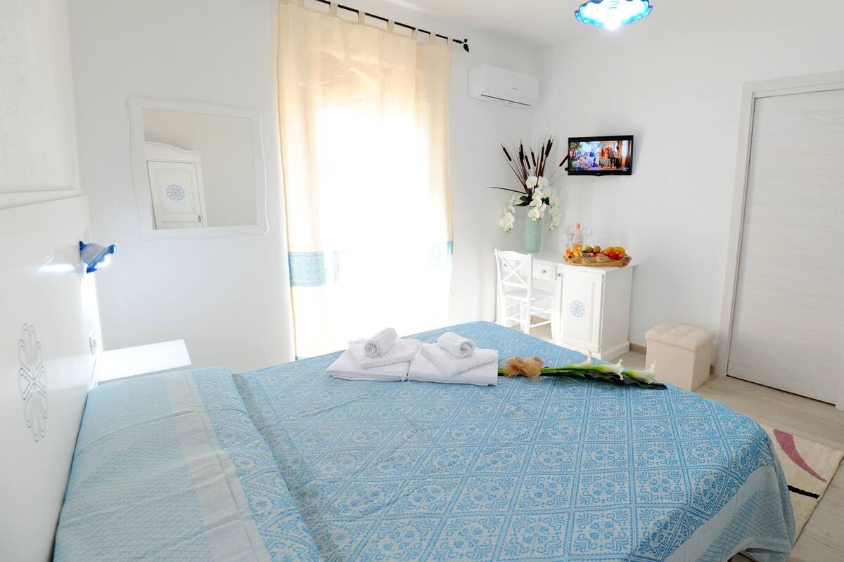 A neatly arranged bedroom with a bed, a small white cabinet, a television, and a window with curtains.