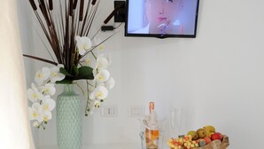 A white desk with a vase of white orchids, a bowl of fruit, and a television on the wall. - INAH Boutique Rooms (Dorgali)