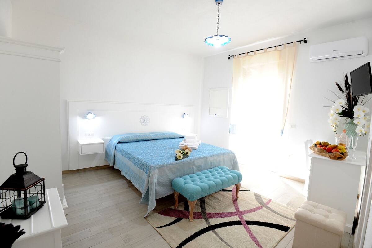 A bedroom with a bed, a bench, a nightstand, a lamp, and a wall-mounted TV.