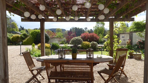 An outdoor wooden pergola with a round table and chairs, surrounded by gravel. - Characterful Home in beautiful surroundings of Surrey Hills (Bramley)