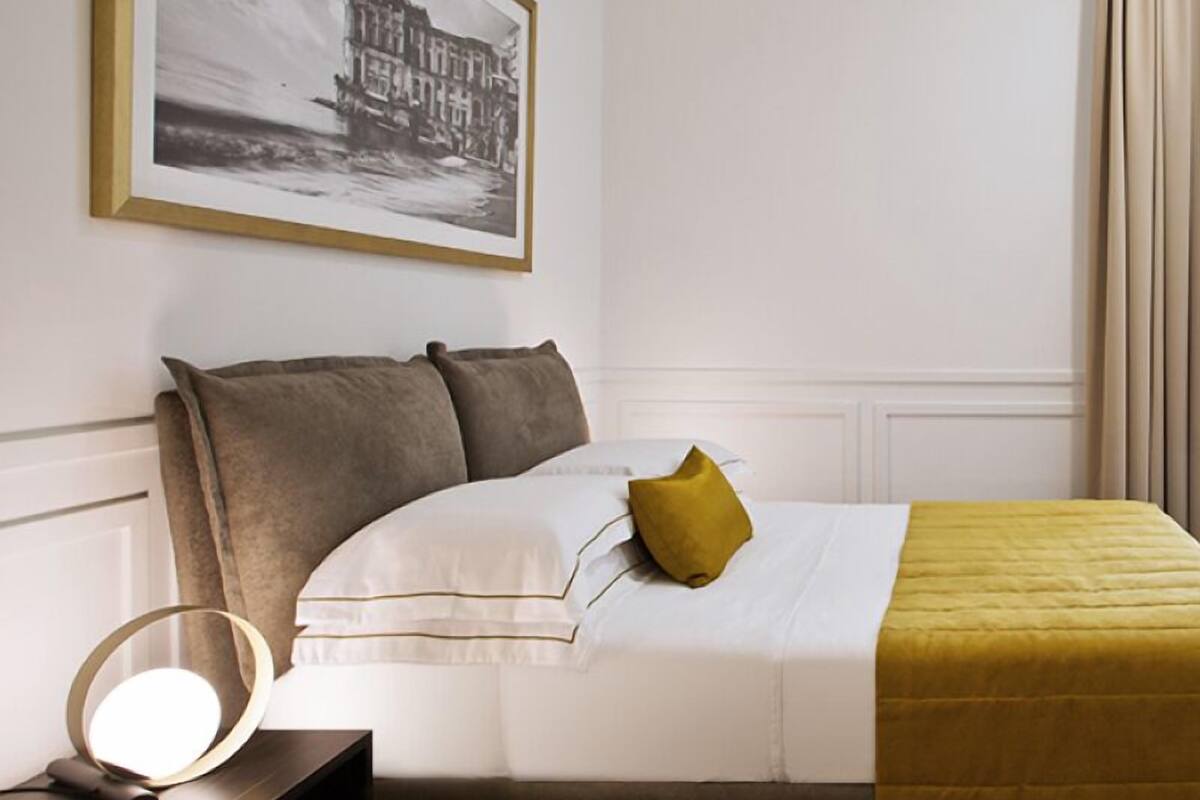 A hotel room with a bed, a bedside table with a lamp, a framed picture on the wall, and a window with curtains.