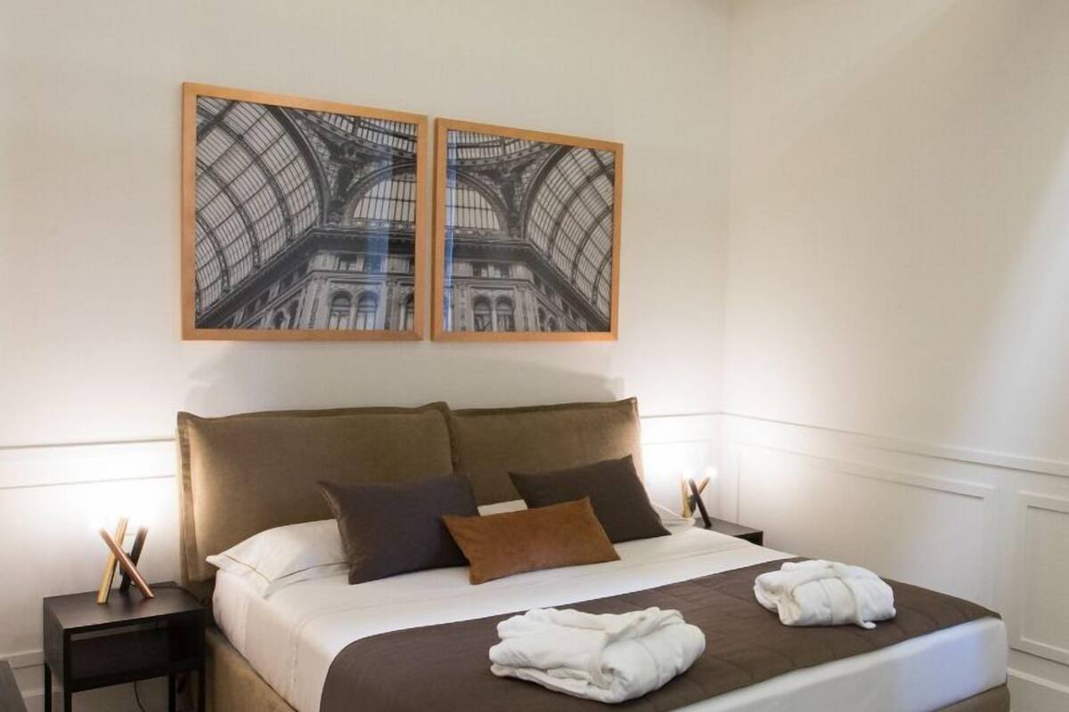 A neatly made bed with a brown headboard, white linens, and two pillows. There are two framed pictures above the bed, a small bedside table with a lamp, and a dark wooden nightstand.