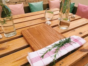 Une table en bois sur laquelle reposent deux bouteilles en verre contenant du romarin, une planche à découper en bois et un tissu orné de motifs de romarin.