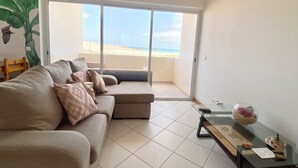 Ein Wohnzimmer mit einer Couch, einem Glastisch und Blick auf den Strand durch eine Schiebetür.
