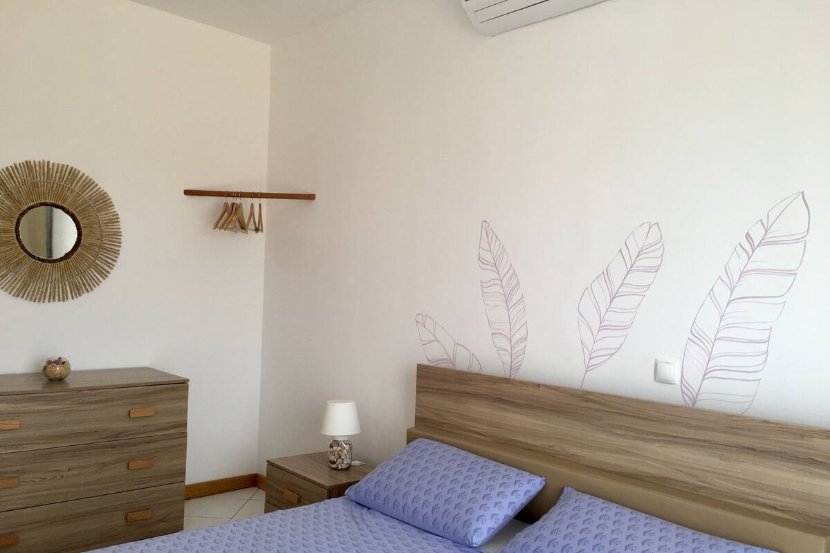 Un dormitorio con cabecera de madera, cama con ropa de cama azul, mesita de noche con lámpara, cómoda, espejo y pared decorada con motivos de plumas.