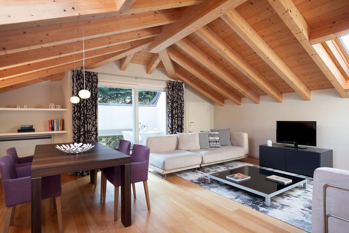 Uma sala de estar moderna com teto de madeira, uma área de jantar com cadeiras roxas, um sofá, uma mesa de centro e uma televisão sobre um móvel preto.