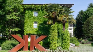 Ein Gebäude mit einem großen 'K'-Schild, umgeben von üppiger Bepflanzung und einem gepflegten Garten.
