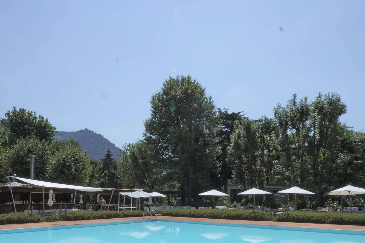 Área de piscina com espreguiçadeiras e guarda-sóis, rodeada por árvores e uma montanha ao fundo.